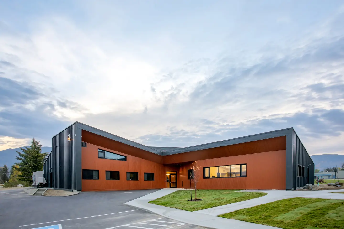 Exterior of The Confluence: a low timber and steel building with burnt-orange cladding, grass and mountains behind it